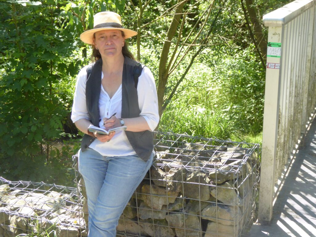 Eine Frau mit Sonnenhut (die Autorin) lehnt im Schatten an einer Mauer unter Bäumen und notiert etwas in einem kleinen Notizbuch.
